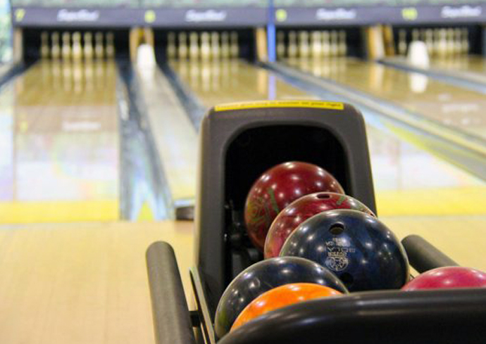 Colorful bowling balls arranged in a ball return rack at a bowling alley, with polished wooden lanes and white pins visible in the background.