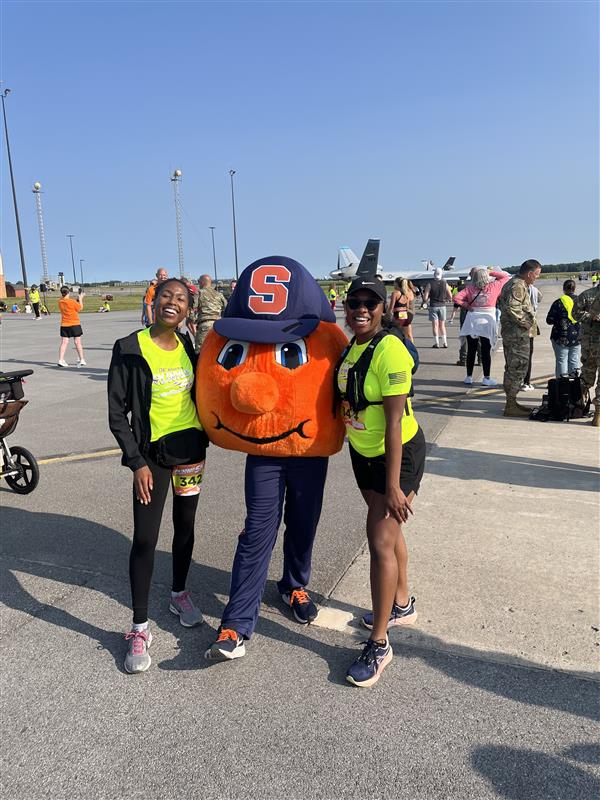 Two women in bright yellow safety vests pose with Otto the Orange, Syracuse University's mascot, on an airport tarmac with military aircraft and personnel visible in the background.