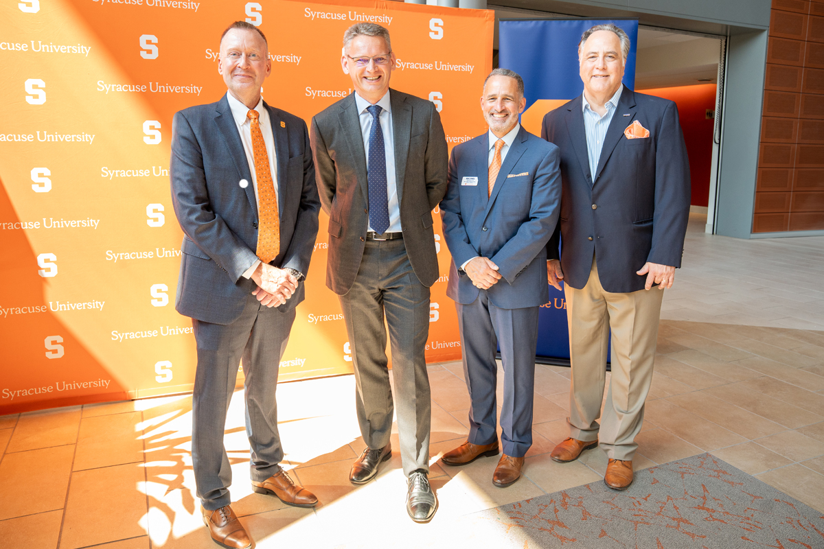 Four men in business suits posing together in front of a Syracuse University branded backdrop. The men are dressed professionally, with one wearing an orange tie that matches the university's colors. 