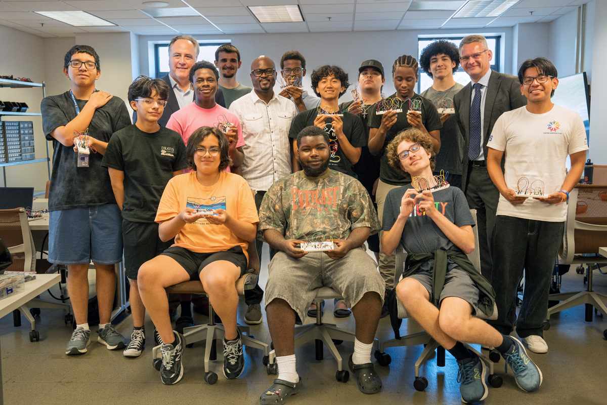 A diverse group of approximately 16 people posing together in what appears to be an office or classroom setting. The group includes students and adults of various ages and ethnicities, with some seated in the front row and others standing behind them. Many are holding what look like small trophies or awards.