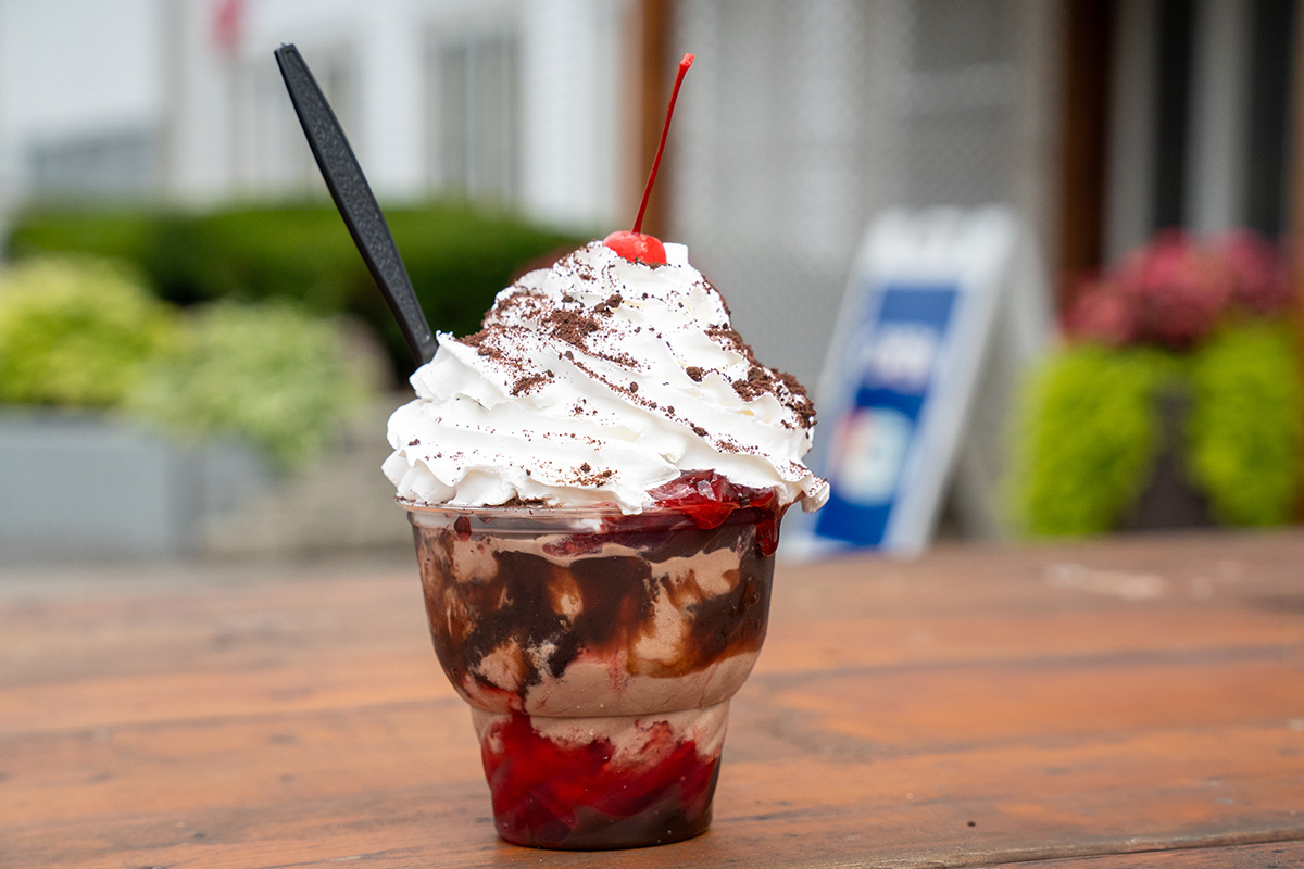 An ice cream sundae in a clear plastic cup with ice cream swirled with chocolate syrup and strawberry sauce, topped with whipped cream, chocolate shavings, and a maraschino cherry, with a black plastic spoon inserted; outdoor background with greenery and signage.