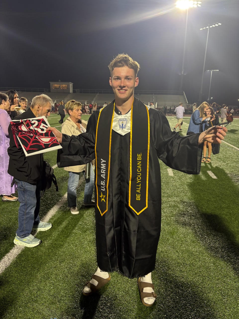 Young man in a black graduation gown with a U.S. Army sash and people on a field in the background