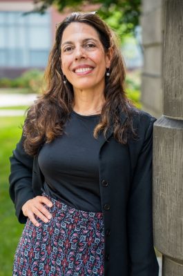 Person with long, wavy hair standing outdoors next to a stone wall, wearing a black top, black jacket, and patterned skirt; one hand on hip and the other resting on the wall. The background includes green bushes and a building with large windows.