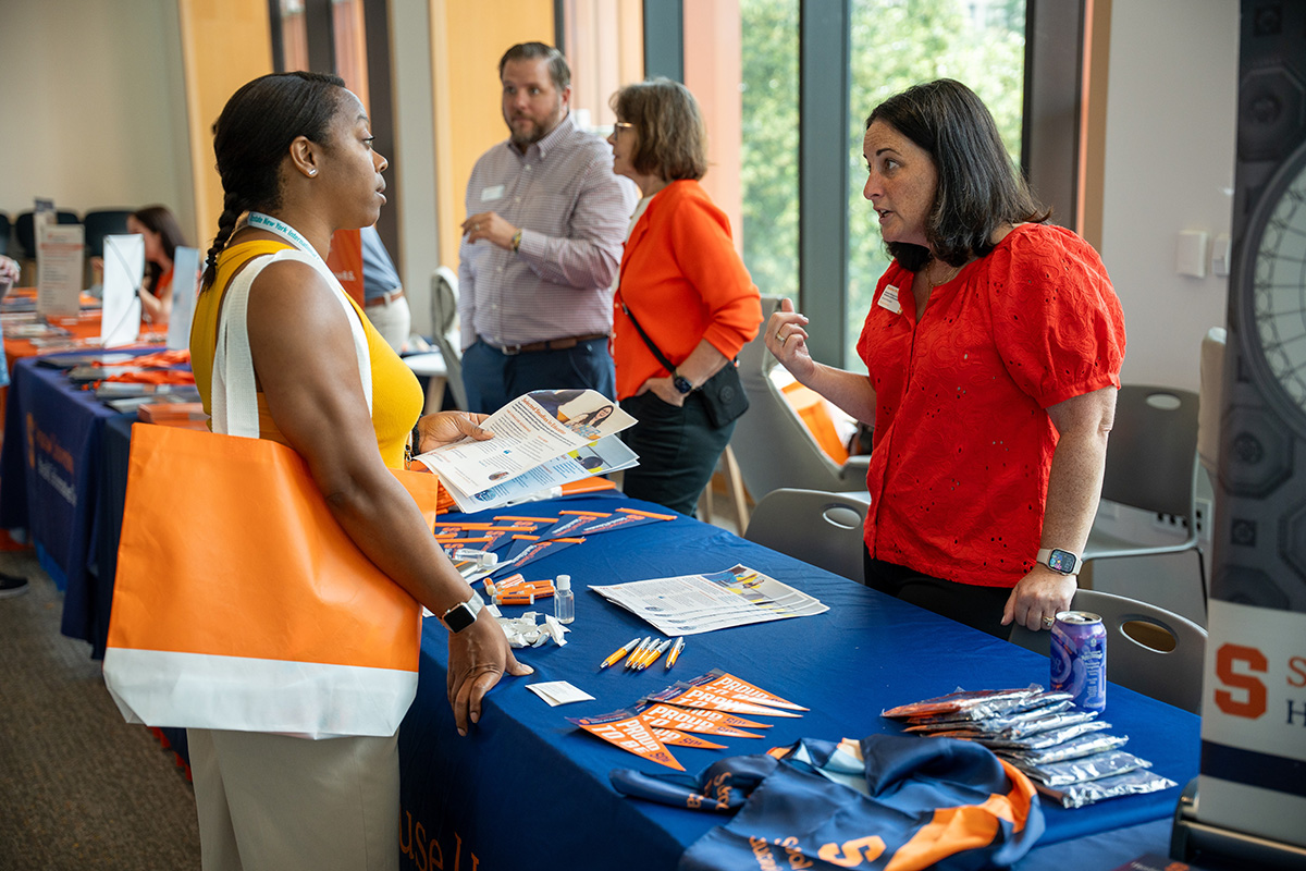 A group of people interacting at an admissions event in Schine Student Center. Tables with blue tablecloths display brochures, pens, and other promotional items. One person with an orange tote bag is holding a brochure, while another in a red shirt gestures toward the table. Additional tables and attendees are visible in the background, with large windows letting in natural light.