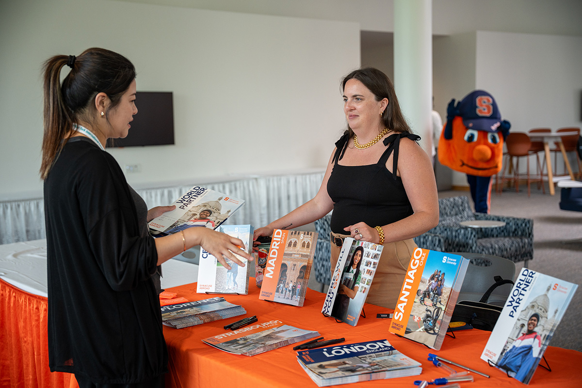 Two people stand at a table displaying print materials promoting Syracuse Abroad, including pamphlets labeled 'MADRID,' 'SANTIAGO,' and 'LONDON.' In the background, the Otto the Orange mascot is visible.
