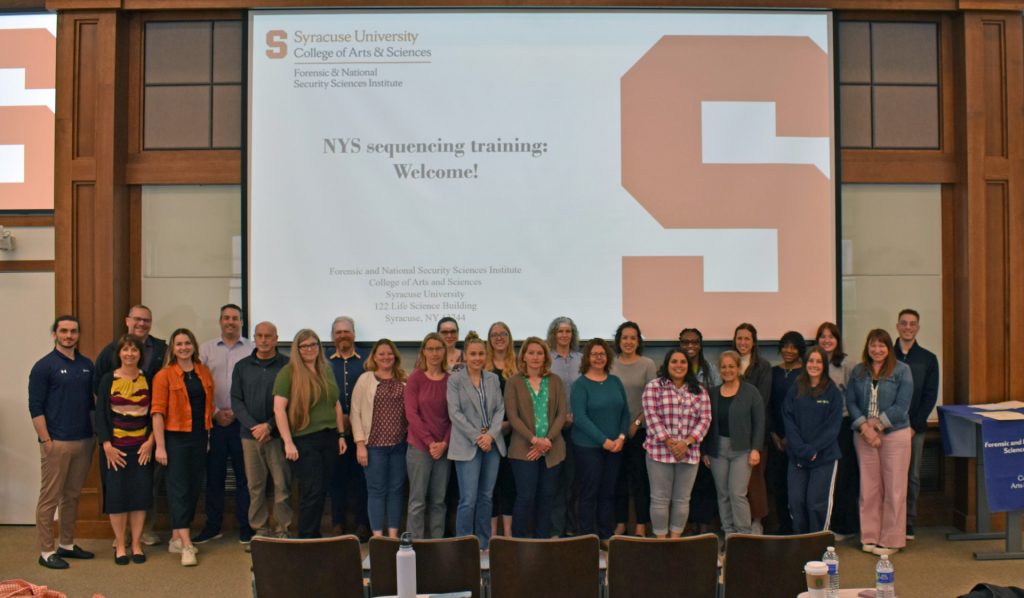 People standing in front of a projection screen at Syracuse University displaying 'NYS sequencing training: Welcome!'"