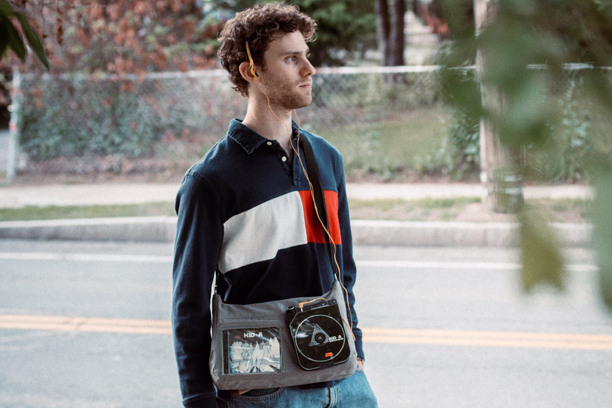 A person standing on the side of a street wearing a navy blue, white, and red color-blocked long-sleeve shirt, with a gray shoulder bag featuring two patches—one with the Kid A album cover by Radiohead and another circular patch with 'KID A' and an abstract design.