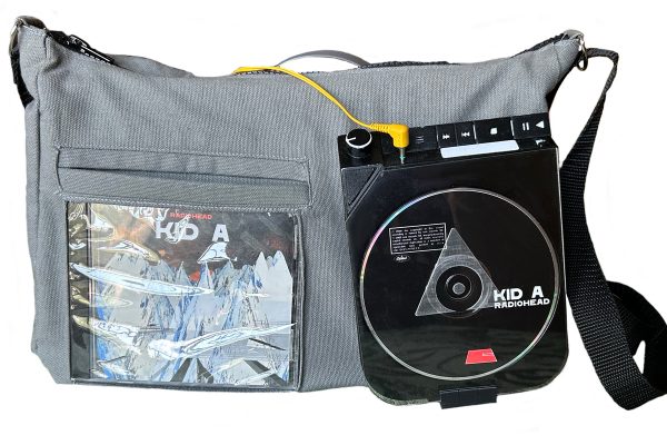 Gray shoulder bag with a black strap, featuring a transparent front pocket displaying Radiohead's 'Kid A' album cover. A portable CD player is attached to the front of the bag, with the 'Kid A' CD inside and a yellow headphone cable plugged in.