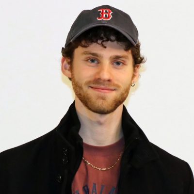 Person wearing a dark baseball cap with a red 'B' logo, a black coat, and a red shirt underneath. The background is plain white.