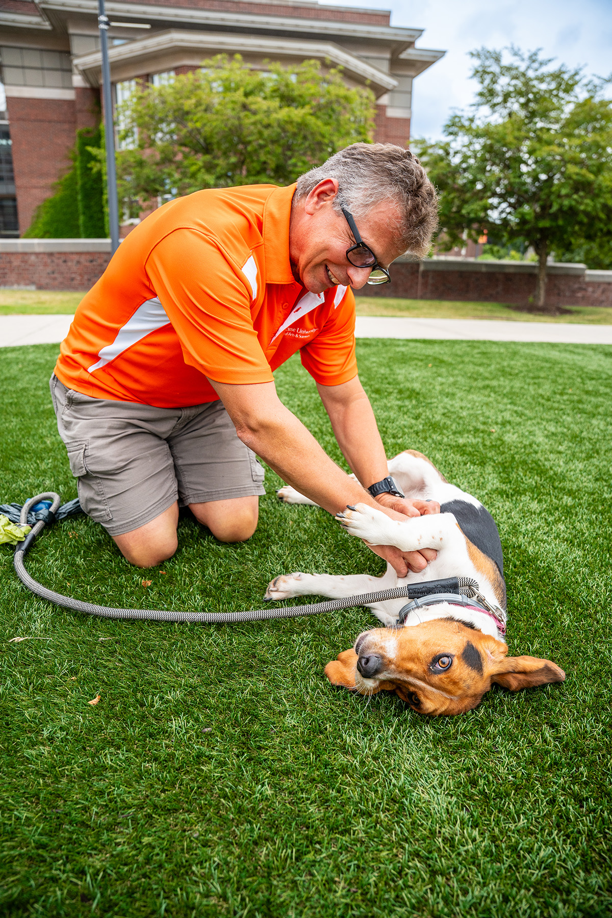 a person in an orange shirt kneeling on grass, gently holding the front paws of a relaxed basset hound lying on its back, with a brick building and trees in the background.