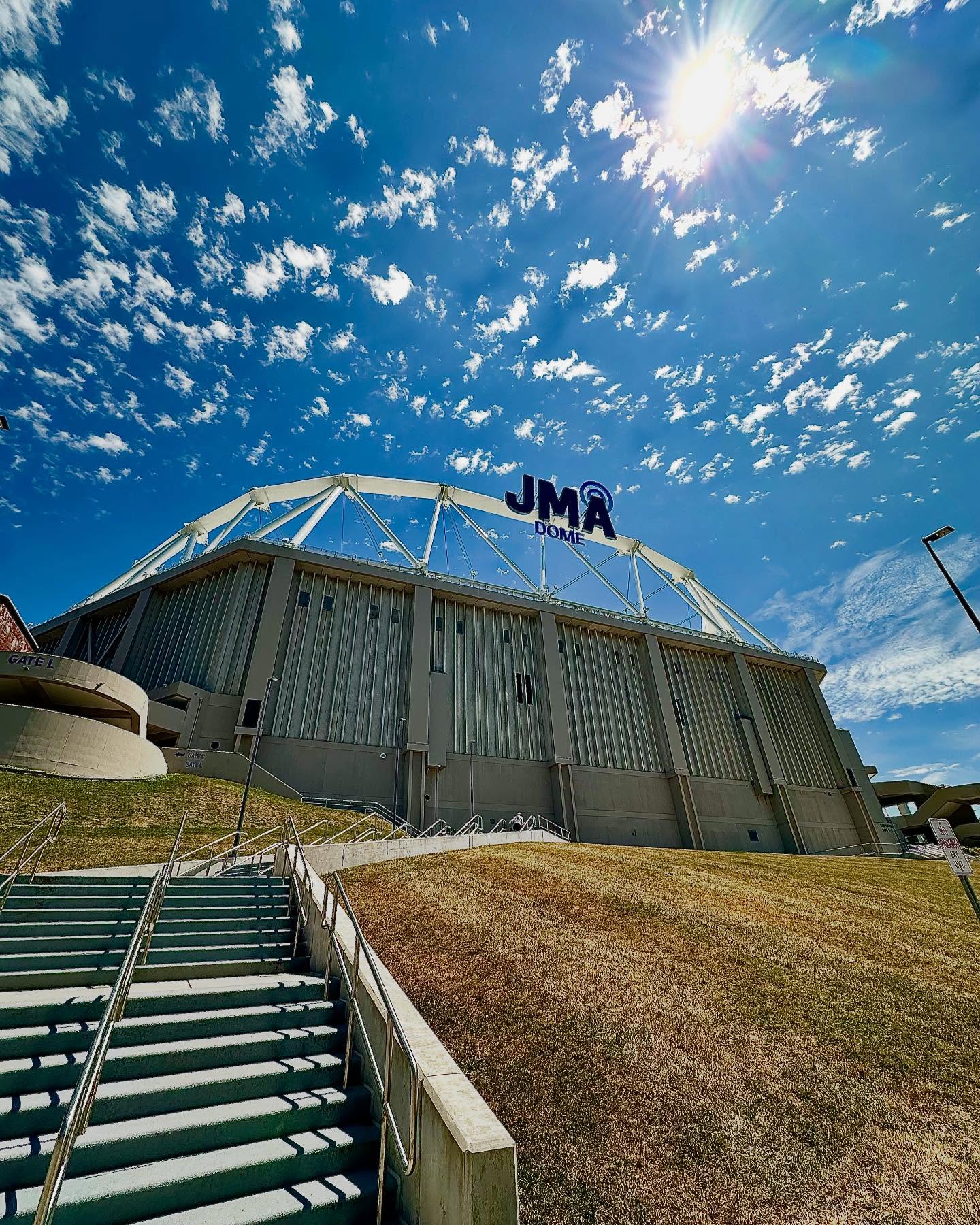 A dramatic low-angle view of the JMA Dome shot against a brilliant blue sky with scattered white clouds and bright sunlight creating lens flares. The modern arena features a distinctive white steel truss roof structure and gray exterior walls. In the foreground, concrete steps with metal handrails lead up a grassy hillside toward the building. The 'JMA DOME' signage is prominently displayed on the facility's exterior.