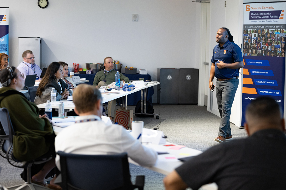 A man in a navy Syracuse University polo shirt presents to a classroom of seated attendees, with a banner for the D'Aniello Institute for Veterans & Military Families visible in the background.