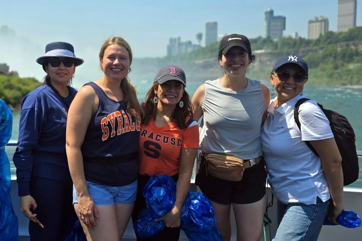 Five individuals stand on a boat with a scenic backdrop of water and tall buildings. They are casually dressed and holding blue plastic ponchos while in front of Niagara Falls.