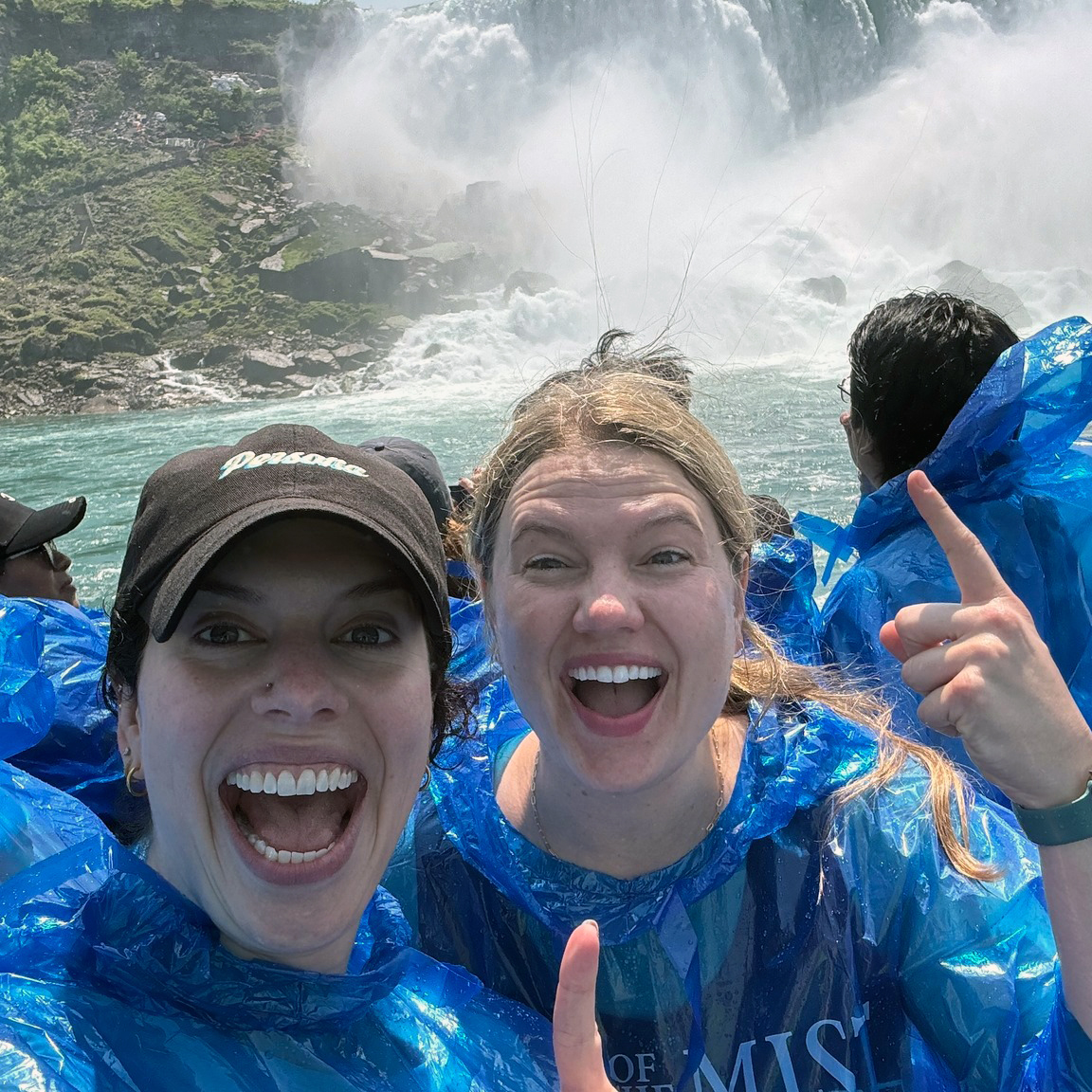 Two people in blue rain ponchos stand near a large, powerful waterfall, with mist rising in the background and turbulent water below. One person is pointing upward with their index finger. 