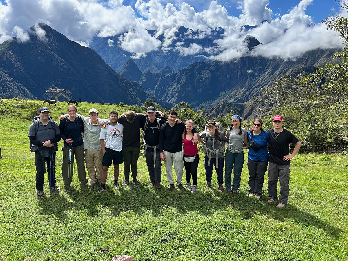 12 people standing arm-in-arm on a grassy field with mountains and clouds in the background during an immersion trip to Peru