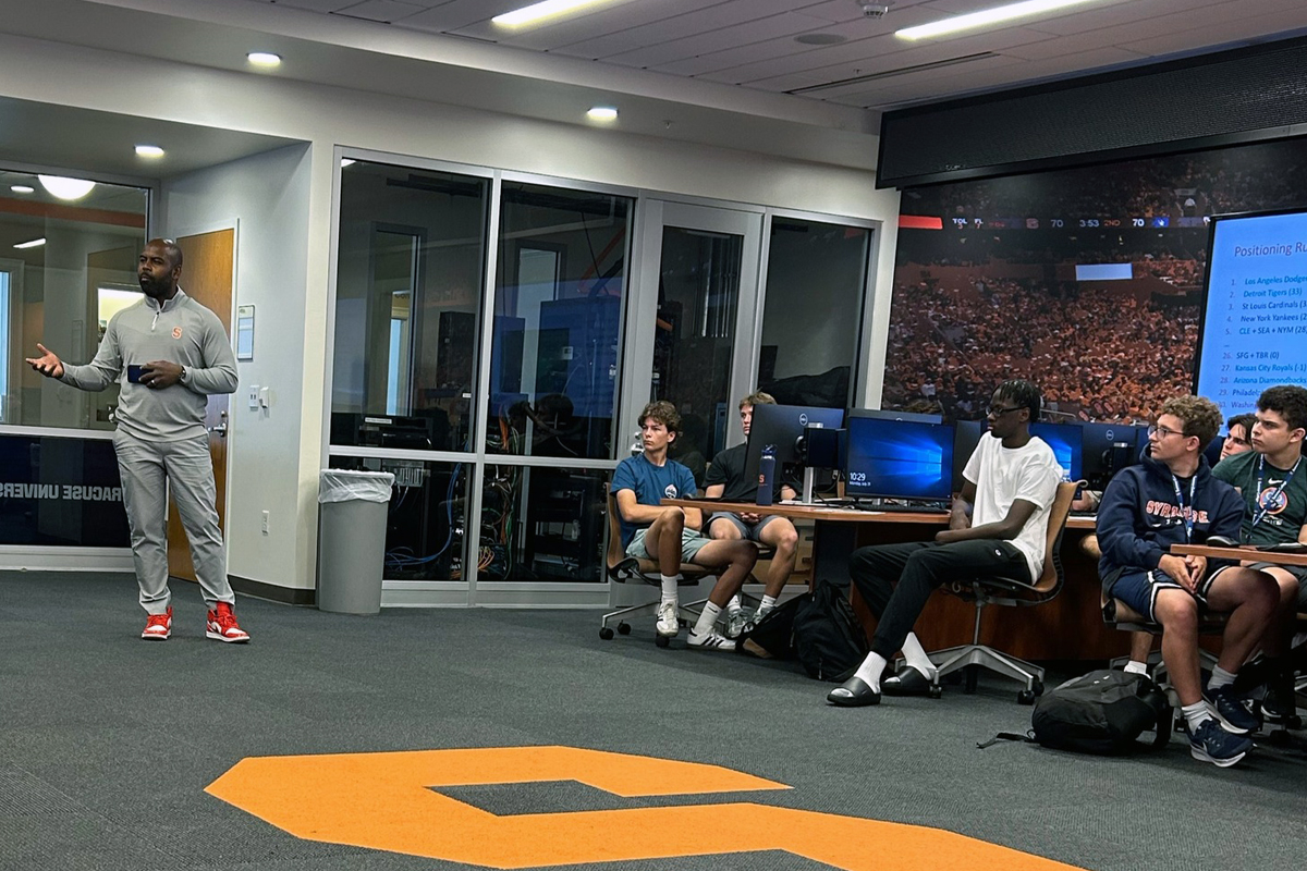 A man in a gray Syracuse University tracksuit and red sneakers stands presenting to a group of young men seated in office chairs in what appears to be a modern sports facility or training room. The presenter is gesturing while speaking to approximately 6-7 young people, likely student-athletes, who are listening attentively. Behind them is a large wall-mounted screen displaying what appears to be a basketball arena filled with fans. The room features floor-to-ceiling windows, modern lighting, and a large orange 'S' logo (Syracuse University's symbol) prominently displayed on the gray carpeted floor.
