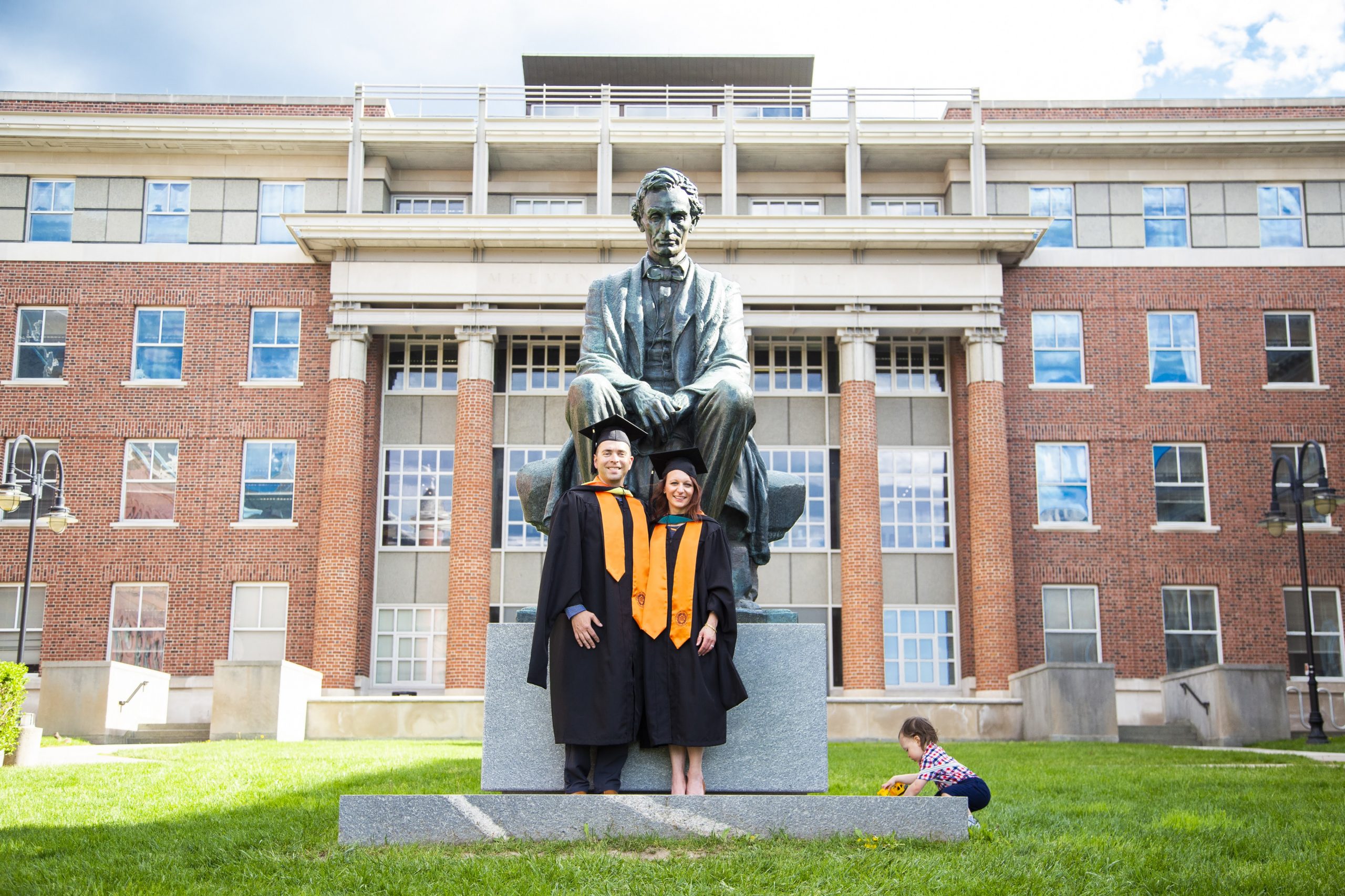 A statue of a seated Abraham Lincoln is positioned in front of a large brick building with many windows. Two people in graduation gowns and caps stand on the base of the statue. A child is seen playing on the grass to the right of the statue.