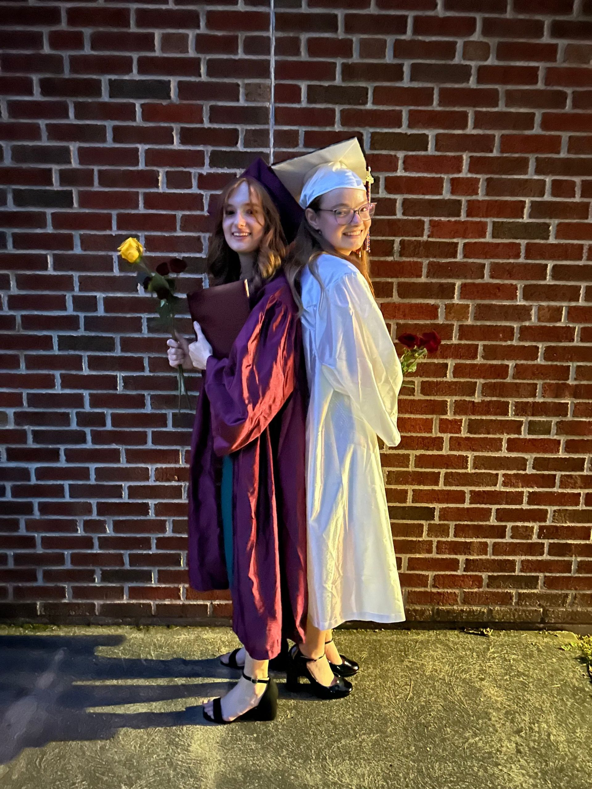 Two young women, one in red graduation attire holding a yellow rose and one in white graduation attire holding red flowers.