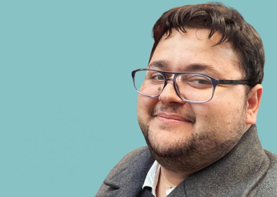 A smiling person with dark wavy hair and glasses wearing a gray sweater or cardigan over a white collared shirt, photographed against a soft teal or mint green background. He has a beard and appears to be looking slightly off-camera with a pleasant, friendly expression.