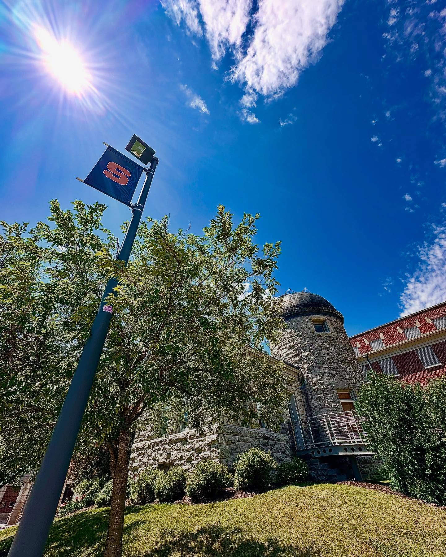 A wide-angle view looking up at a sunny blue sky with white clouds, featuring a lamp post with a Syracuse University 'S' logo sign in the foreground. Behind it stands a distinctive stone building with a cylindrical tower topped by a dark dome, characteristic of historic university architecture. The building is surrounded by lush green trees and landscaping, with a well-maintained lawn in the foreground. A red brick building is partially visible in the background. The bright sun creates a lens flare effect in the upper left portion of the image.