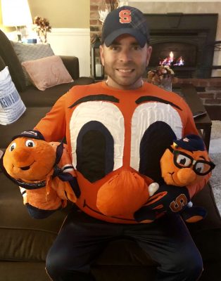 A person sits on a couch wearing an orange shirt featuring a large cartoon face resembling Otto the Orange, the Syracuse University mascot. They hold two Otto the Orange plush toys, one in each hand, and wear a blue cap with an orange block S. Behind them is a lit fireplace and various home decor items.
