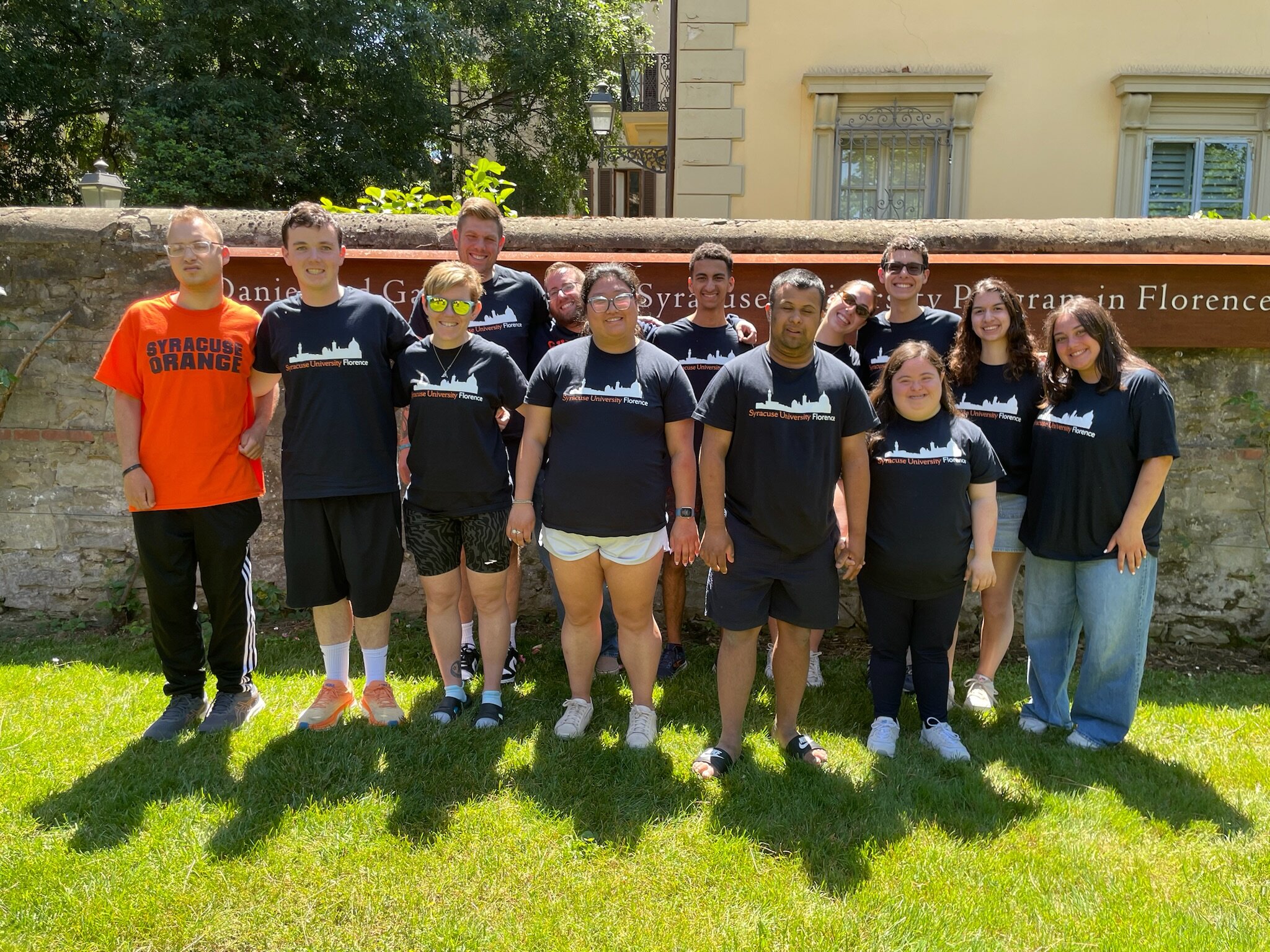 A group of 13 people stands on a grassy area in front of a stone wall with a sign that reads 'Daniel and Gayle D'Aniello Syracuse University Program in Florence." Many of the individuals wear matching black t-shirts with white and orange text that says "Syracuse University Florence," while one person on the far left wears an orange t-shirt that says "Syracuse Orange." Behind them is a building with windows and surrounding greenery.