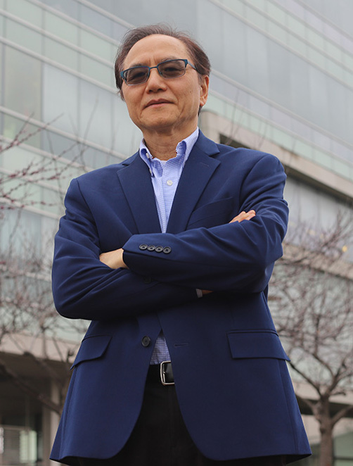 Person standing with arms crossed in front of a modern glass building, wearing a blue blazer, light blue shirt, and black pants. The background includes leafless trees, suggesting autumn or winter.
