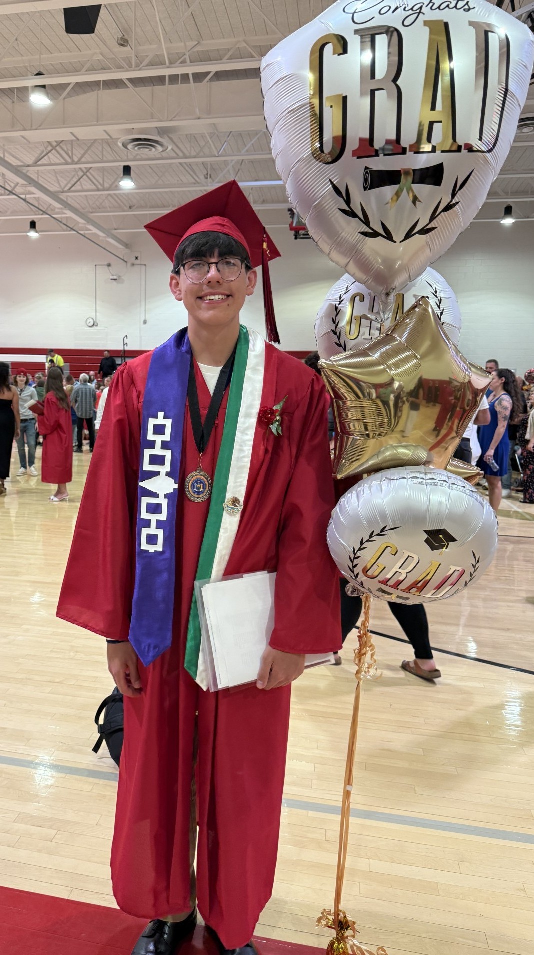 Young man in red graduation gown and cap with Native American sash, standing by a bouquet of balloons in a gymnasium
