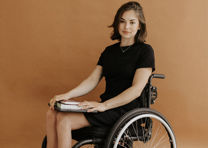 Young woman with shoulder-length brown hair wearing a black dress sits in a wheelchair, holding a book and smiling at the camera against a warm beige background.