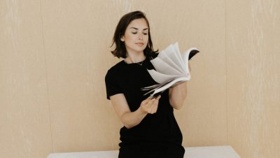 Young person in a black short-sleeved dress reading an open book with pages fanned out, standing against a neutral beige background