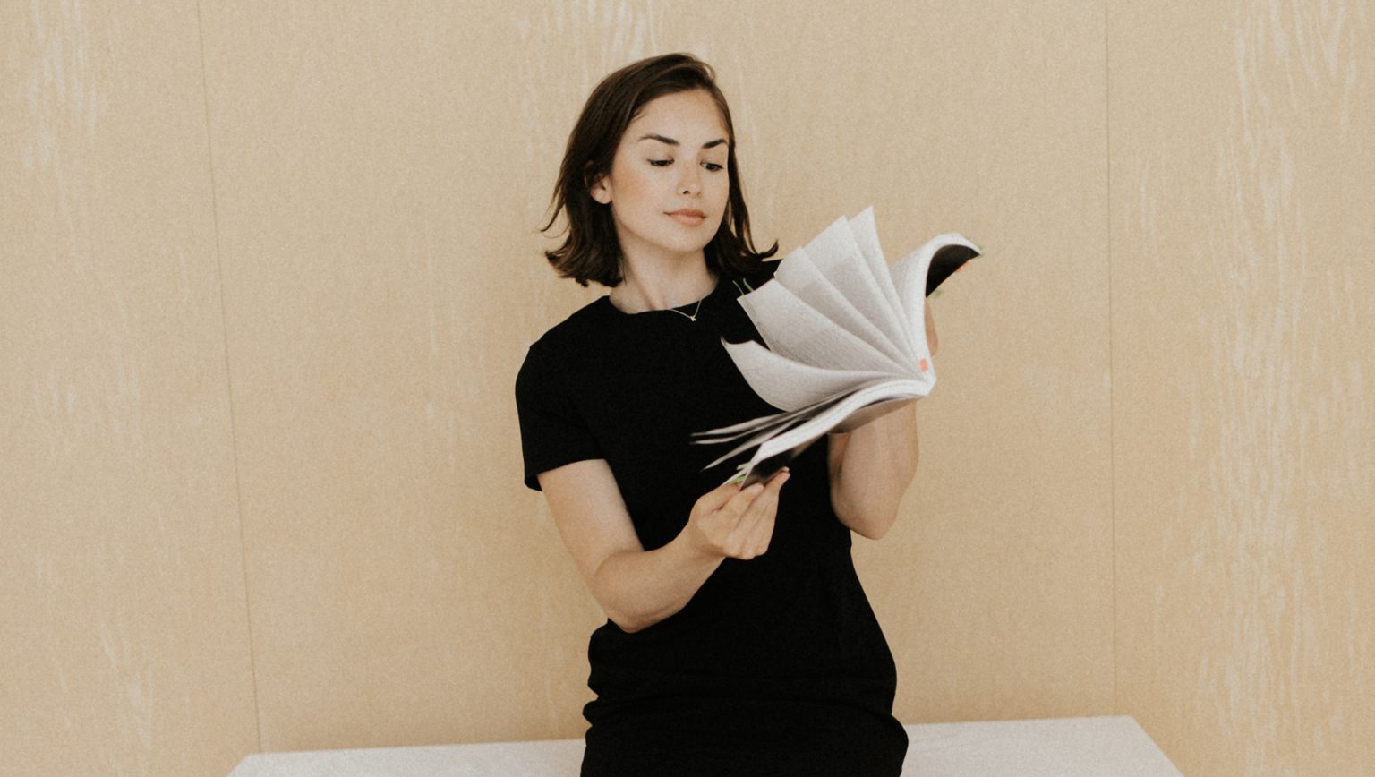 Young person in a black short-sleeved dress reading an open book with pages fanned out, standing against a neutral beige background.
