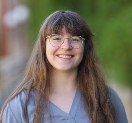 A person with long, straight brown hair and bangs smiles warmly at the camera. They are wearing round, wire-rimmed glasses and a light blue or periwinkle colored top with a V-neck. The photo appears to be taken outdoors with a soft, blurred green background suggesting trees or foliage.