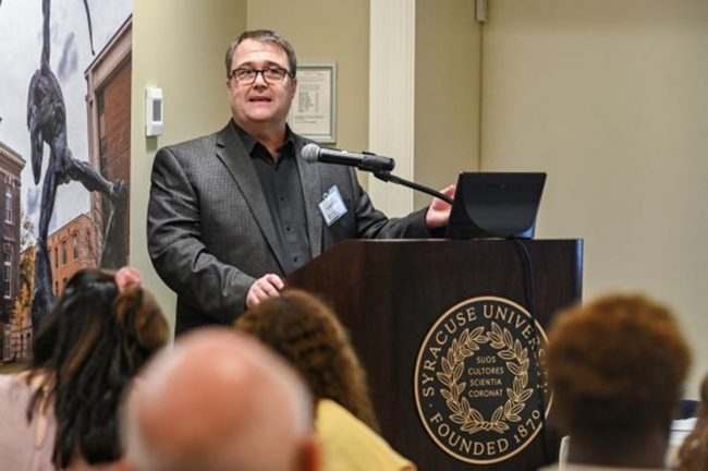 A person stands at a Syracuse University lectern addressing an event audience