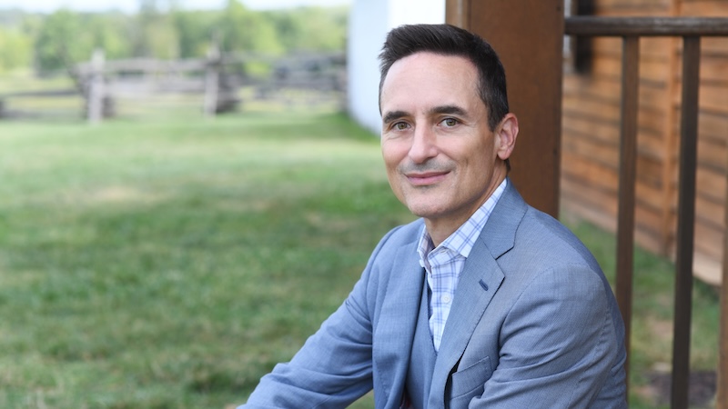 Person in a light blue suit and checkered shirt seated outdoors near a wooden structure, with grassy area and trees in the background.