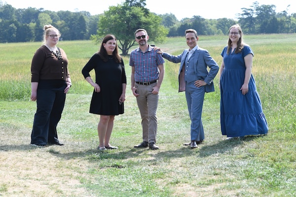 Group of five people standing in a grassy field with trees in the background on a sunny day, dressed in casual and formal attire
