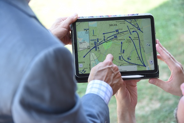Two people reviewing a tablet displaying a detailed map with blue route markings, outdoors in a grassy area.