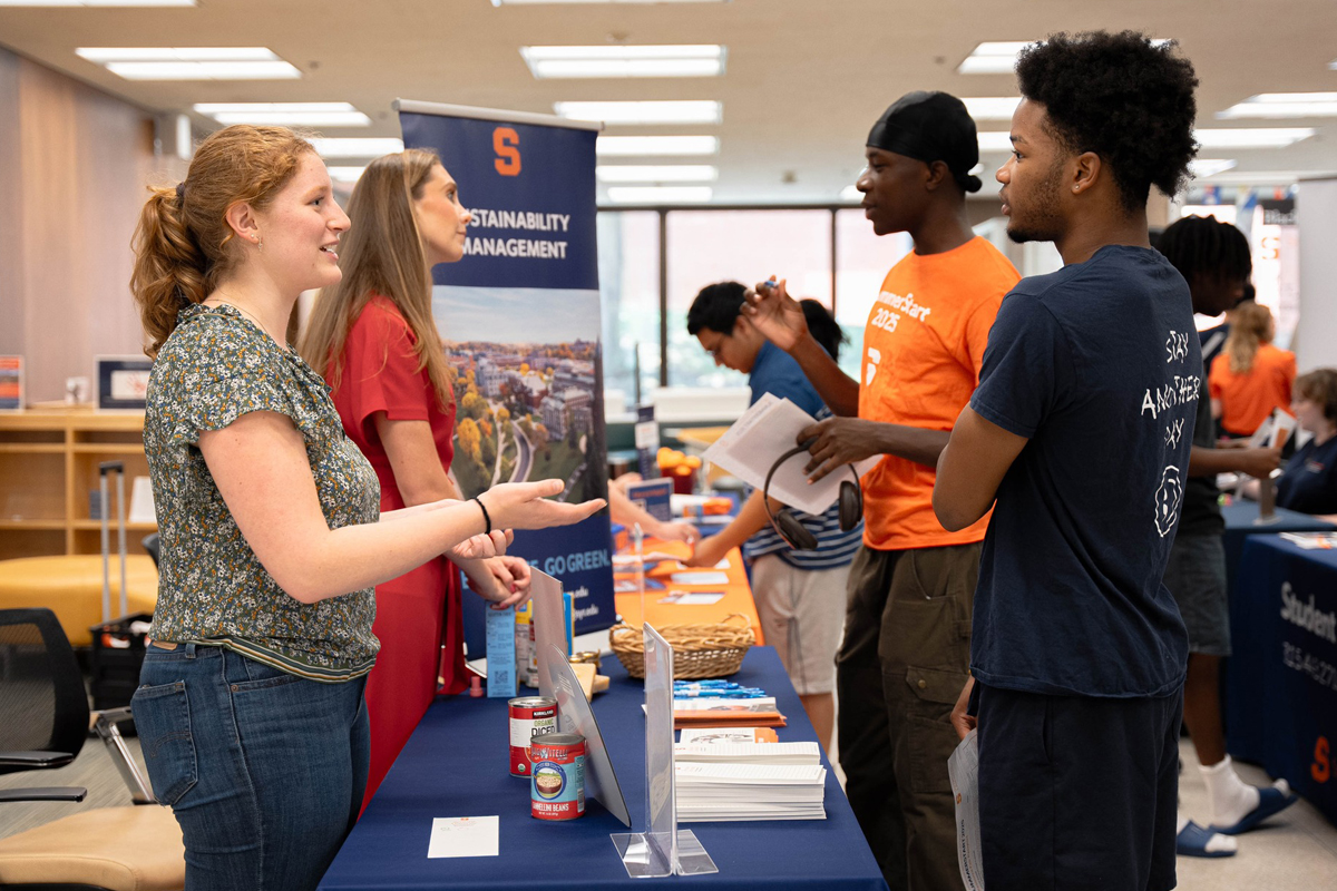 Two people at a Syracuse University Sustainability Management information booth speak with two male students, one wearing an orange shirt and one in a navy shirt.