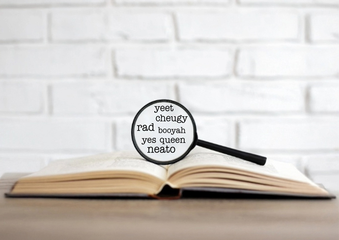 Open book on wooden surface with magnifying glass highlighting slang words like 'yeet' and 'cheugy'; white brick wall in background