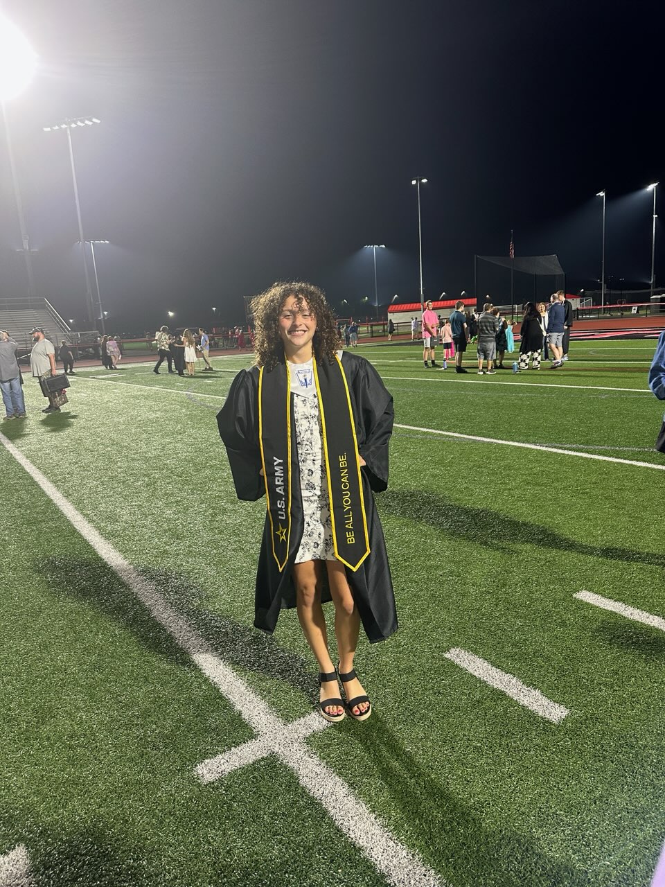 Young woman in black graduation gown with U.S. Army sash standing on a field