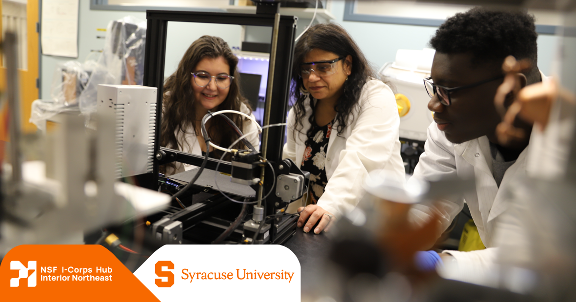 Three researchers in white lab coats and safety glasses working together in a laboratory setting. Two women are positioned on the left side of the image, with one wearing purple-framed glasses, both smiling as they observe scientific equipment. A third person is on the right side, focused on hands-on work with laboratory instruments. The scene shows various pieces of scientific equipment and apparatus on lab benches. In the bottom left corner are logos for the NSF I-Corps Hub Interior Northeast and Syracuse University.