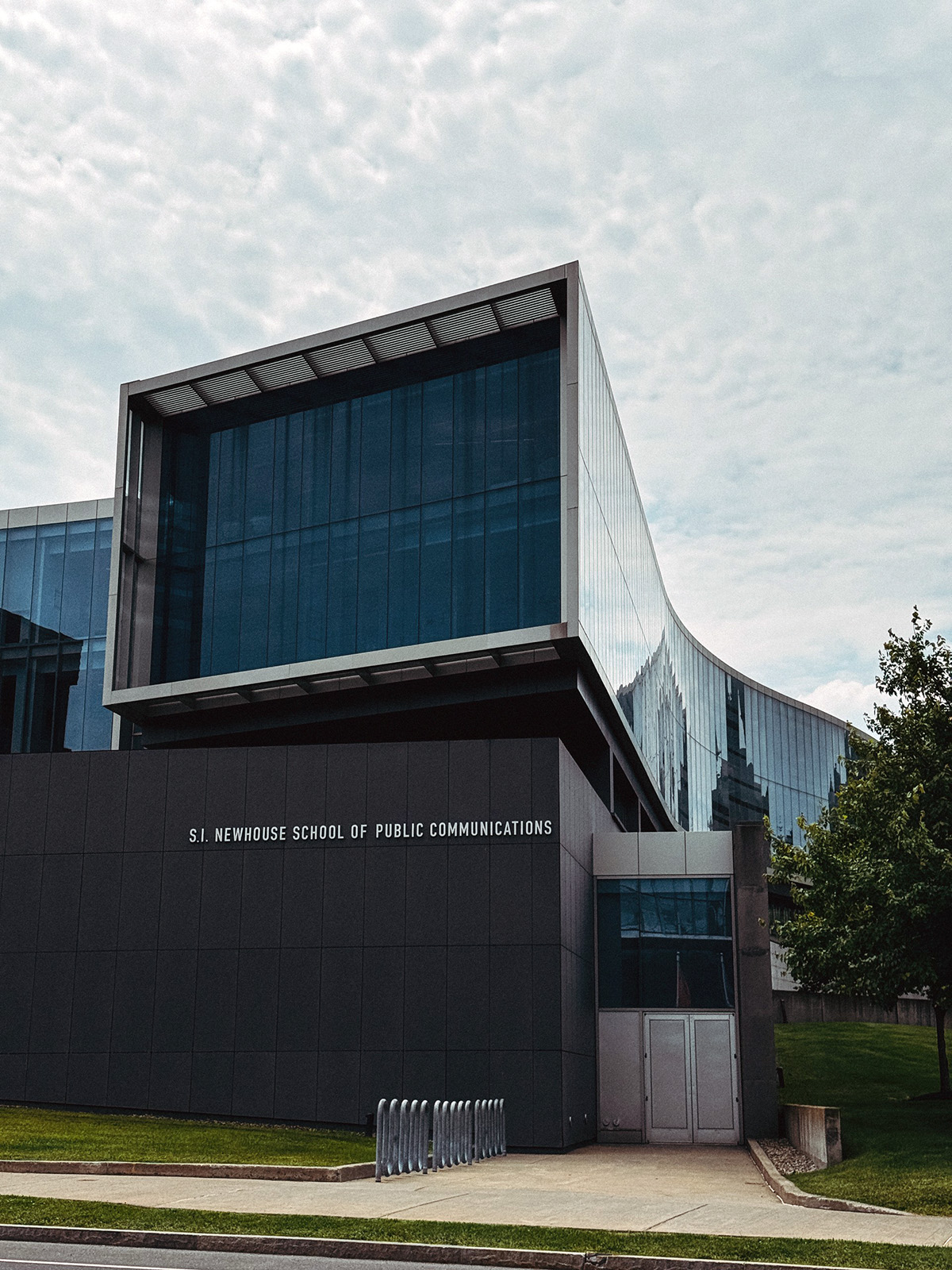 Exterior of the S.I. Newhouse School of Public Communications with modern glass architecture and visible signage under a cloudy sky.