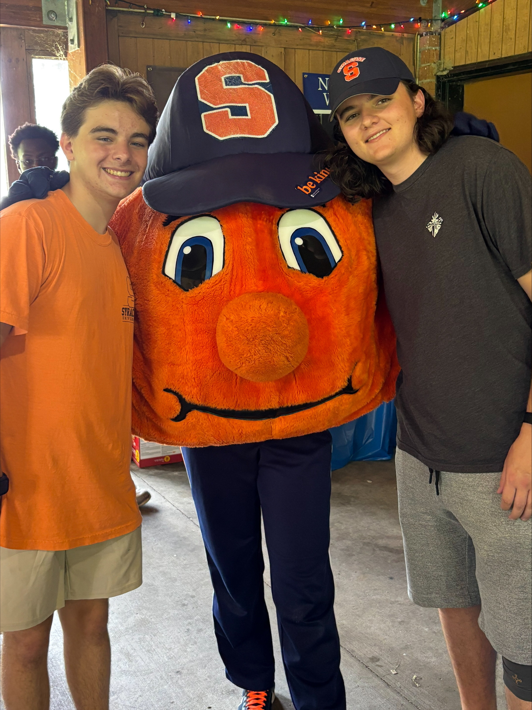 Otto the Orange flanked by two young men in t-shirts and shorts. One is wearing a Syracuse cap