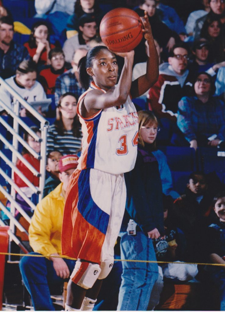 Basketball player wearing a white jersey with red and blue accents and the number 34, captured mid-jump shot while holding a Spalding basketball above their head. The background includes spectators seated in bleachers.
