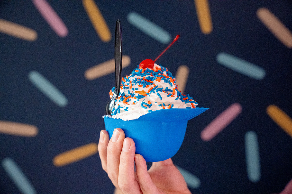 Hand holding a plastic blue hat "bowl" of ice cream topped with whipped cream, blue and orange sprinkles, and a cherry, with a black plastic spoon inserted; background features a dark blue wall with a colorful sprinkle pattern.