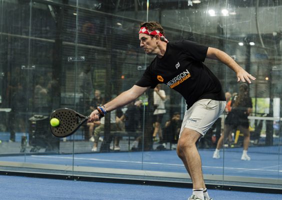 A person playing padel on an indoor court. They are wearing a black shirt, light-colored shorts and a red headband, and are in the act of hitting a yellow ball with a padel racket. Behind them, spectators are seated and watching the game.