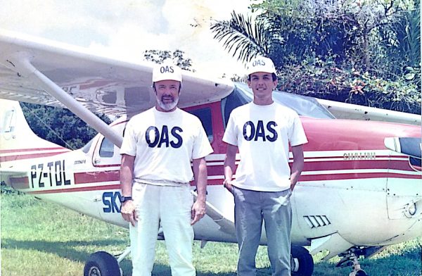 Two individuals wearing white t-shirts and caps labeled 'OAS' stand in front of a small white aircraft with red stripes and the registration number PZ-TOL. The word 'SKYLANE' is visible on the aircraft's side. The background features greenery and palm trees, suggesting a tropical or subtropical setting.