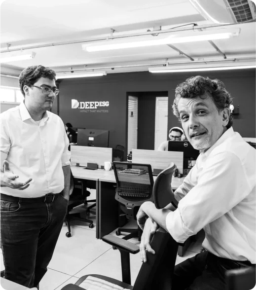 Two individuals engaged in conversation in a modern office setting with multiple workstations, computers, and chairs. 