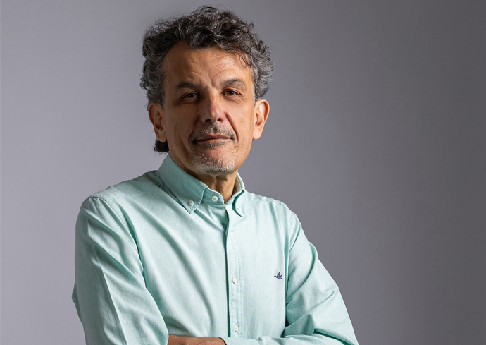 A person with curly dark hair and visible ears, wearing a light green long-sleeved button-up shirt, standing with arms crossed against a plain gray background.
