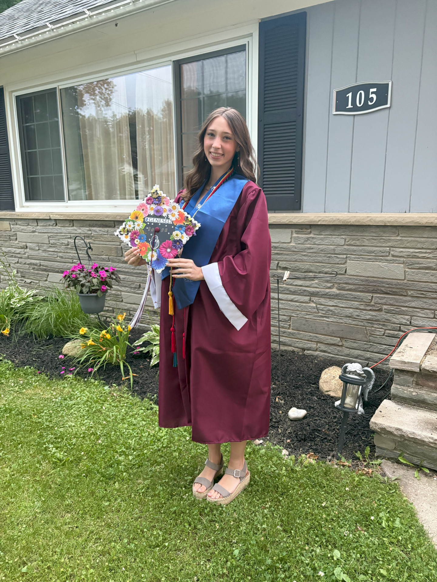 Payton Abbott in red graduation gown standing in front of a house holding a bouquet of flowers