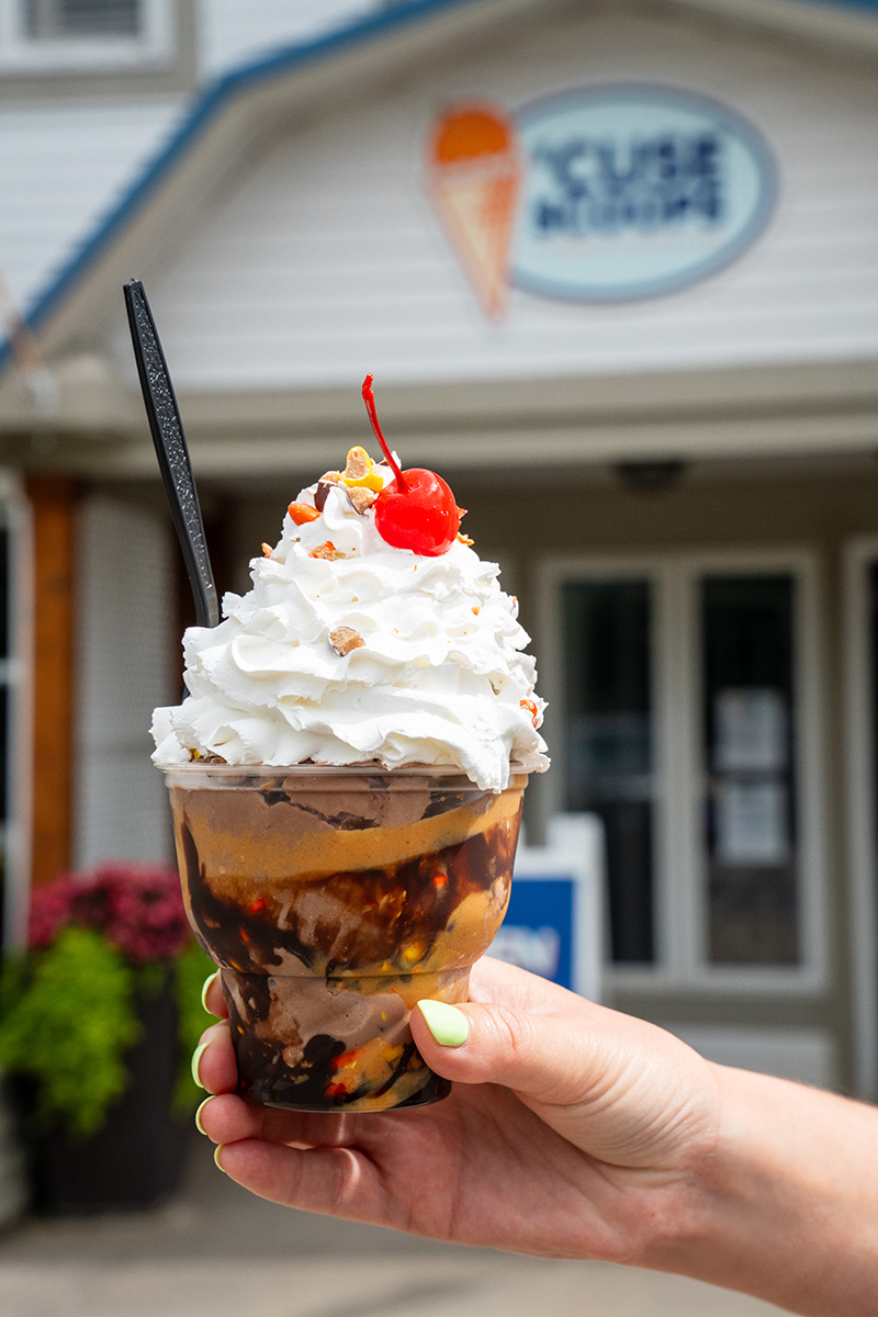  Hand holding a sundae cup with ice cream, chocolate syrup, peanut butter sauce, whipped cream and a cherry; background shows the entrance of an ice cream shop with a 'Cuse Scoops sign and ice cream cone logo.
